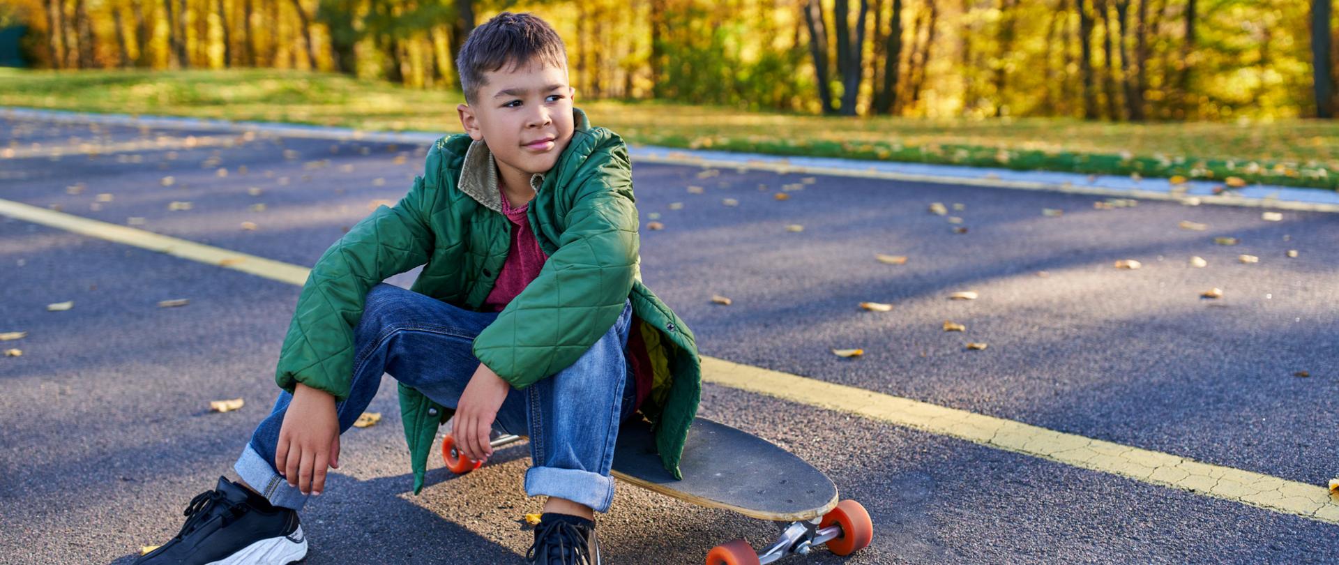 Na zdjęciu jest chłopak siedzący na deskorolce. W tle widać las.