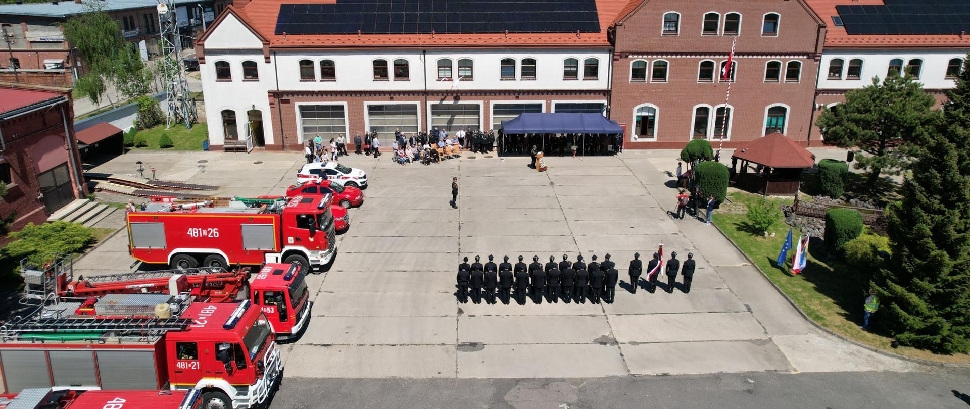 00 Zdjęcie przedstawia strażaków na uroczystym apelu z okazji Dnia Strażaka. Widok z drona z góry, Pol lewej stronie widoczne ustawione w szeregu samochody pożarnicze