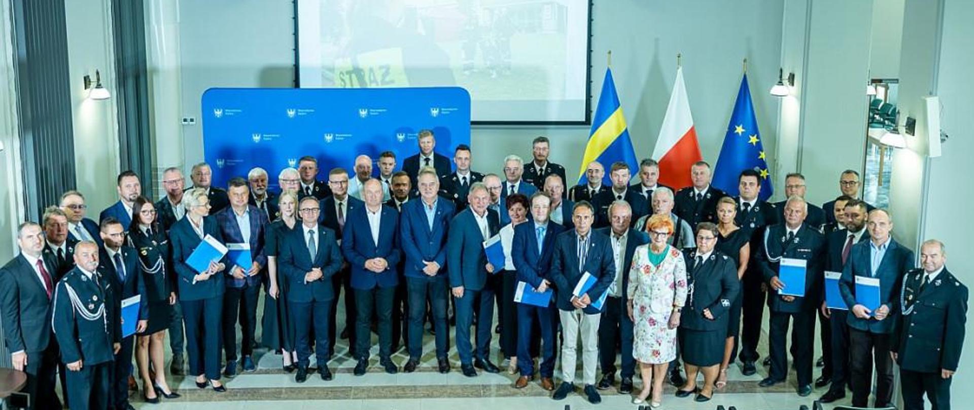 Na zdjęciu widzimy grupę około 30 osób pozujących do zdjęcia w sali Urzędu Marszałkowskiego. Stoją w trzech rzędach. Osoby są ubrane w stroje formalne, a niektóre mają na sobie mundury straży pożarnej.W tle znajduje się duży ekran i niebieska ścianka. Pomieszczenie ma wysokie sufity z żyrandolami, a obok ekranu widoczne są flagi Unii Europejskiej, Polski i woj. śląskiego