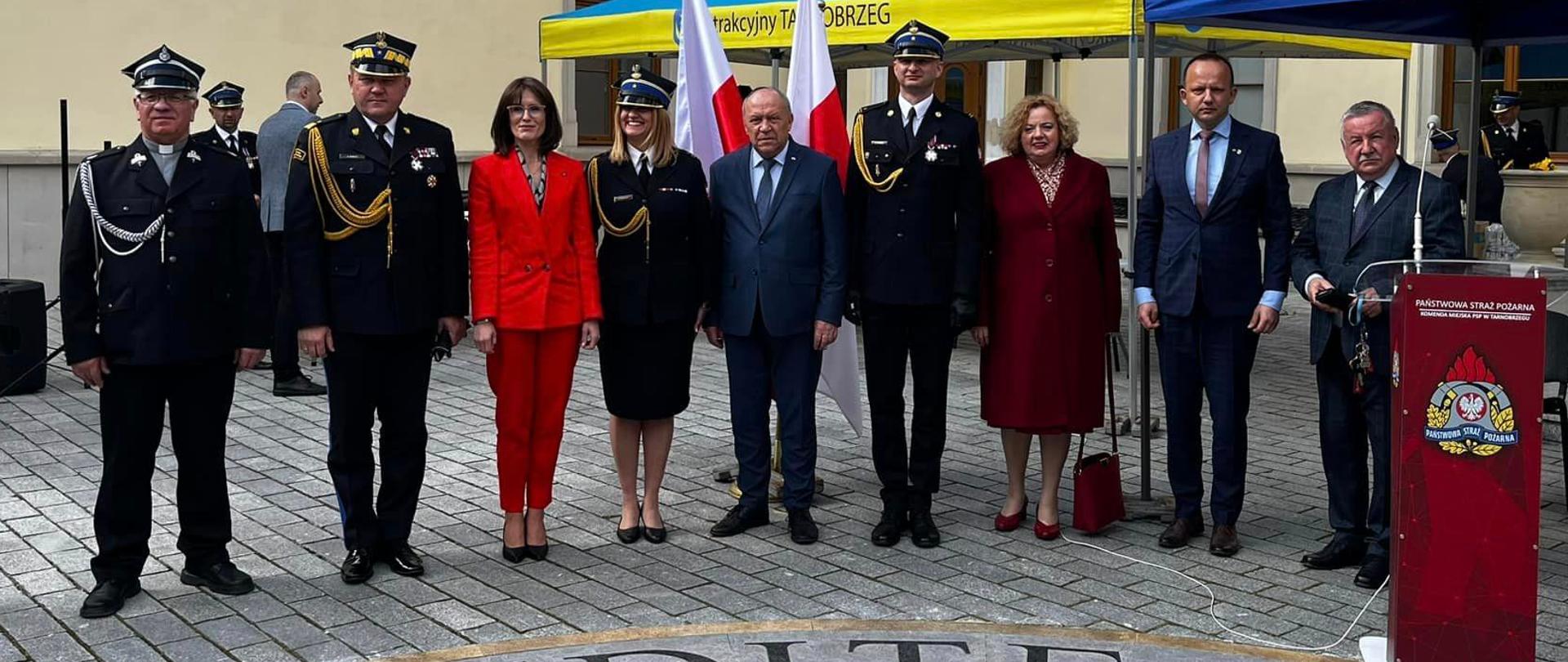 Zdjęcie zrobione na zewnątrz podczas uroczystości. Na zdjęciu, w szeregu stoją zaproszeni na uroczystość goście. Za nimi stoją ustawione namioty na których jest napis Komenda Miejska Państwowej Straży Pożarnej w Tarnobrzegu.