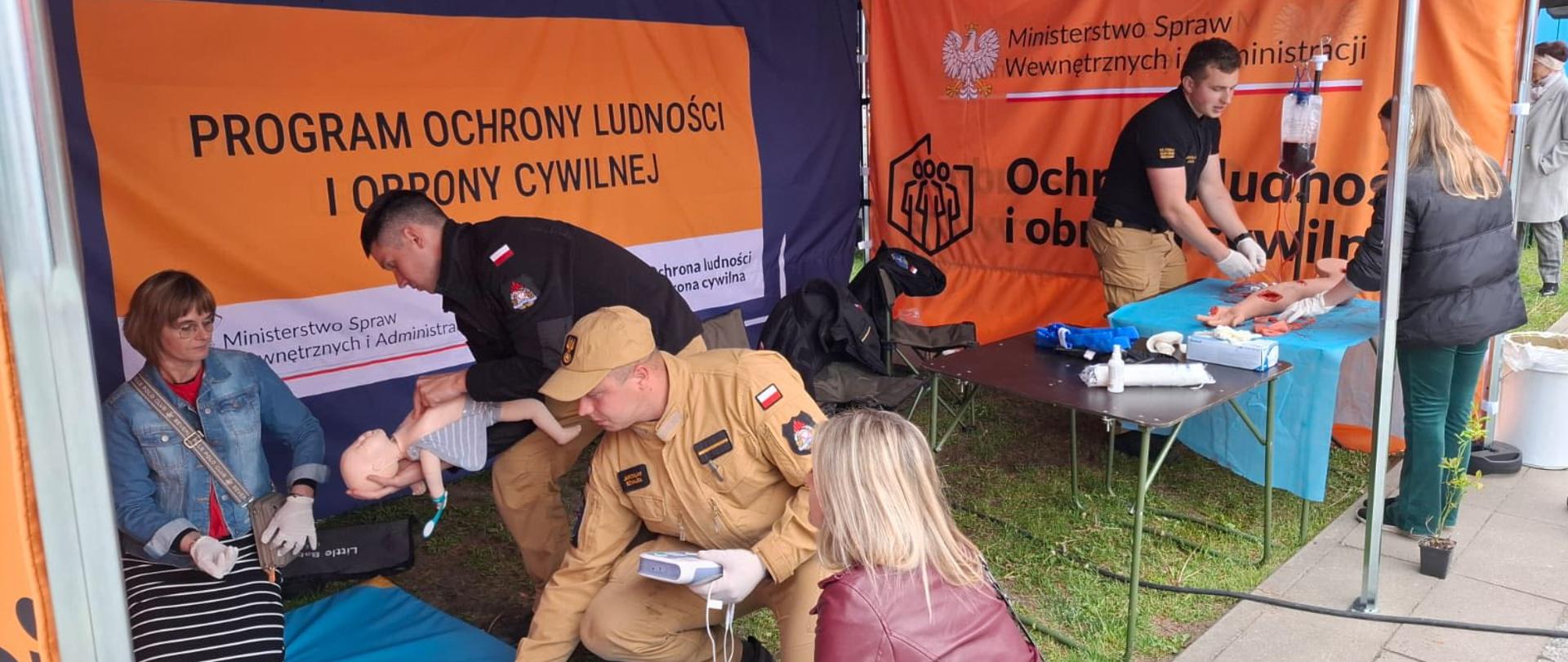 Na zdjęciu widać stoisko edukacyjne pod namiotem, przygotowane w ramach programu „Ochrona Ludności i Obrona Cywilna”. Strażacy prowadzą praktyczne zajęcia z pierwszej pomocy, pokazując uczestnikom, jak prawidłowo reagować w sytuacjach zagrożenia życia.
W centrum i po lewej stronie widać ćwiczenia na fantomie niemowlęcia oraz osobę uczącą się podstawowych czynności ratowniczych. Po prawej stronie odbywa się kolejny pokaz z wykorzystaniem fantomu i materiałów opatrunkowych. Na stole rozłożone są akcesoria medyczne, m.in. opatrunki, rękawiczki i sprzęt do ćwiczeń.
W tle widoczne są banery informacyjne z napisem „Program Ochrony Ludności i Obrony Cywilnej” oraz oznaczeniami Ministerstwa Spraw Wewnętrznych i Administracji. Całość przedstawia praktyczne szkolenie i działania profilaktyczne mające na celu zwiększenie świadomości mieszkańców w zakresie bezpieczeństwa i udzielania pierwszej pomocy.