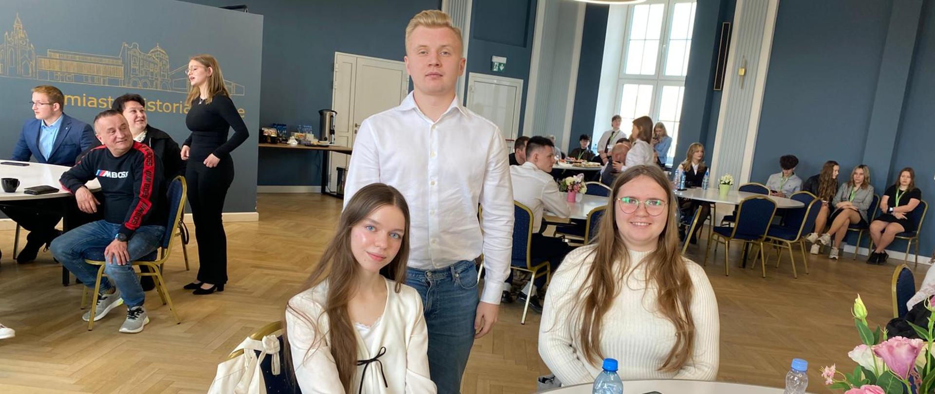 Grupa młodych ludzi pozuje do zdjęcia w dużej, nowoczesnej sali ze ścianami w kolorze ciemnego mosiądzu i parkietem. W centralnej części widać trójkę uśmiechniętych osób siedzących przy stole: kobietę w białej bluzce z kokardką, stojącego za nią mężczyznę w białej koszuli oraz drugą kobietę w swetrze i okularach. Na stole stoją butelki wody, talerzyk z jedzeniem i wazon z kwiatami. W tle, przy innych stołach i na kanapie, widać pozostałych gości spotkania. Nad głowami wiszą duże, okrągłe, ledowe lampy. Po lewej stronie ściany widoczna jest grafika z budynkami i częściowo widoczny napis "miast...storia".