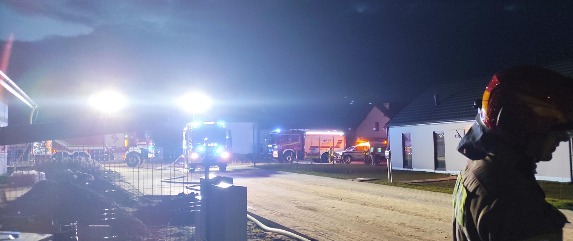 Zdjęcie zrobione nocą. na zdjęciu stoją trzy samochody strażackie na światłach niebieskich ostrzegawczych dodatkowo oświetlając teren przez najaśnice na samochodach. Teren małego osiedla domków jednorodzinnych. Z prawej strony strażak w czerwonym hełmie.