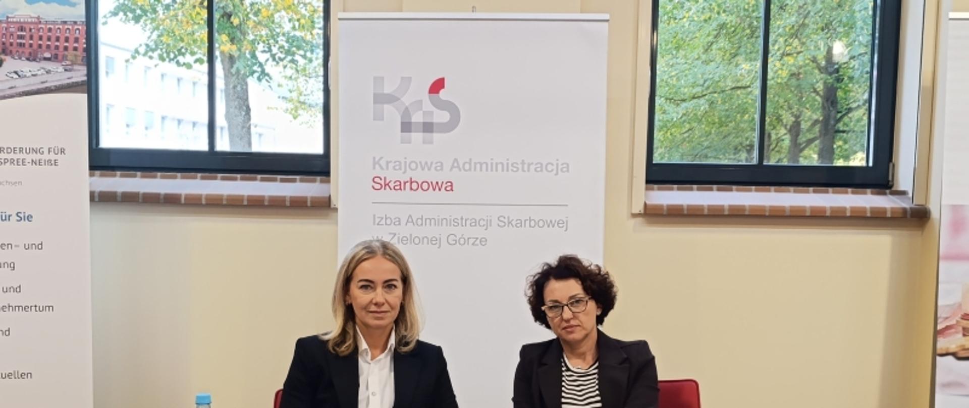 pomieszczenie, za ławką siedzą dwie kobiety, na blacie ławki leżą różne przybory piśmiennicze i ulotki, w tle roll-up z napisem Krajowa Izba Administracji Skarbowej