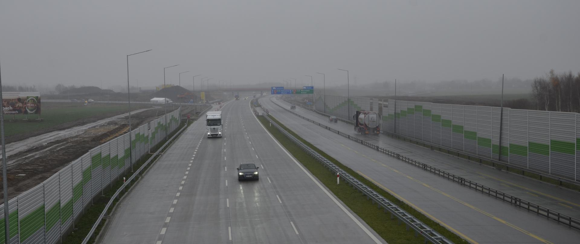 Widoczna skąpana w deszczy wschodni jezdnia autostrady - po lewej stronie, po prawej jezdnia zachodnia. Po obu stronach drogi widoczne są szaro - zielone ekrany akustyczne. W tle widoczny wiadukt łącznicy Warszawa - Katowice.
