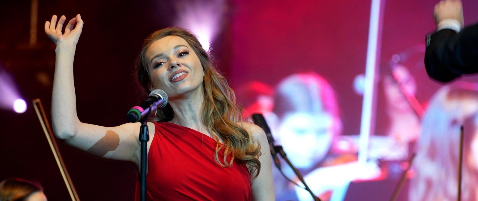 Artystka podczas występu wokalnego. Zdjęcie wykonane od pasa wzwyż. Kobieta ma około 35 lat. Jest ubrana w czerwoną przylegającą sukienkę na jedno ramię. Ma długie brązowe włosy. W tle za nią widać sylwetki muzyków instrumentalistów grających w orkiestrze.