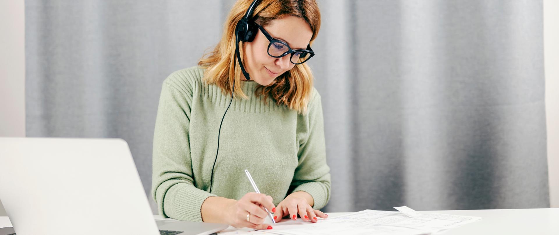 Ruda kobieta w okularach i z słuchawkami na głowie siedzi przy laptopie i notuje