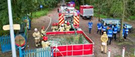 Strażacy przy zbiorniku wodnym zbudowana linia wężowa , pojazdy pożarnicze , obserwatorzy ze Sztabu Zarządzania Kryzysowego Starostwa Powiatowego w Słupcy.