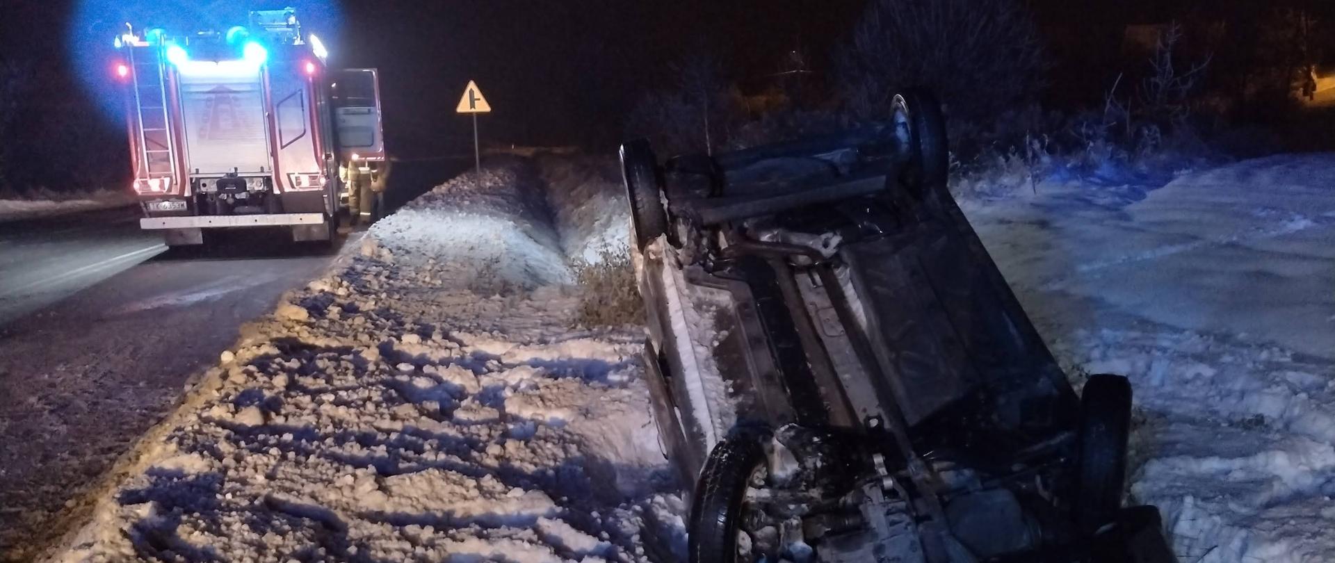 w przydrożnym rowie na dachu leży samochód osobowy. W tle samochód gaśniczy na włączonych światłach niebieskich. Aura zimowa, śnieżna, pora nocna.