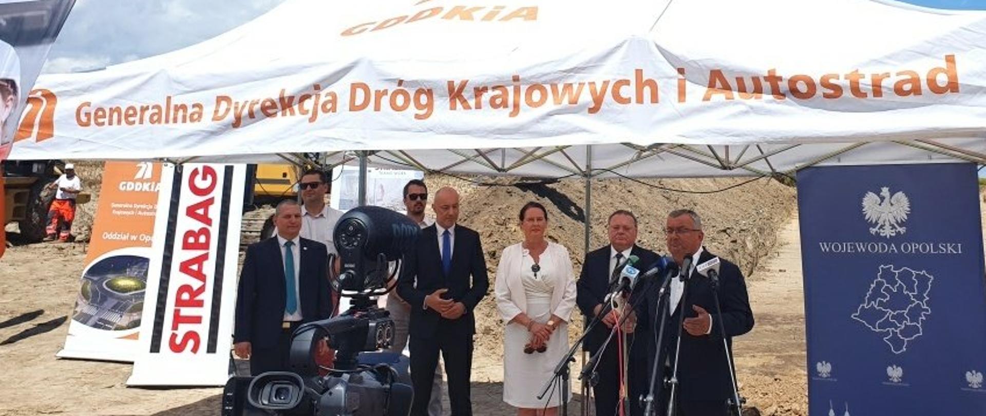Na zdjęciu widać grupę ludzi, która stoi pod namiotem. Jedna z osób stoi przy mikrofonach. W ich tle widać prace budowlane. 