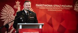 Komendant powiatowy przemawia do obecnych na sali otwierając uroczystość, w tle baner państwowej straży pożarnej
