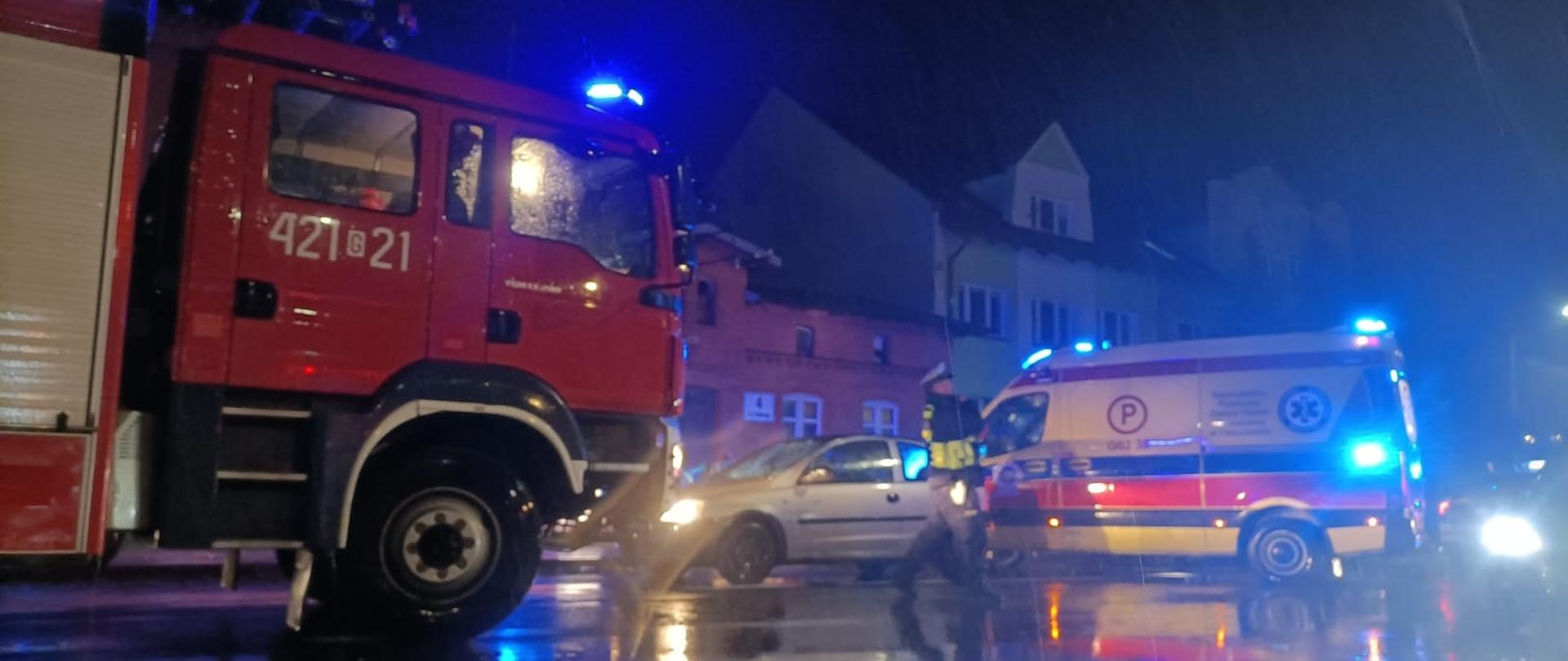 Zdjęcie przedstawia samochód pożarniczy, samochód osobowy i karetkę stojące na ulicy Stefana Batorego w Człuchowie. Przed samochodami przechodzi policjant, w tle widać budynki.