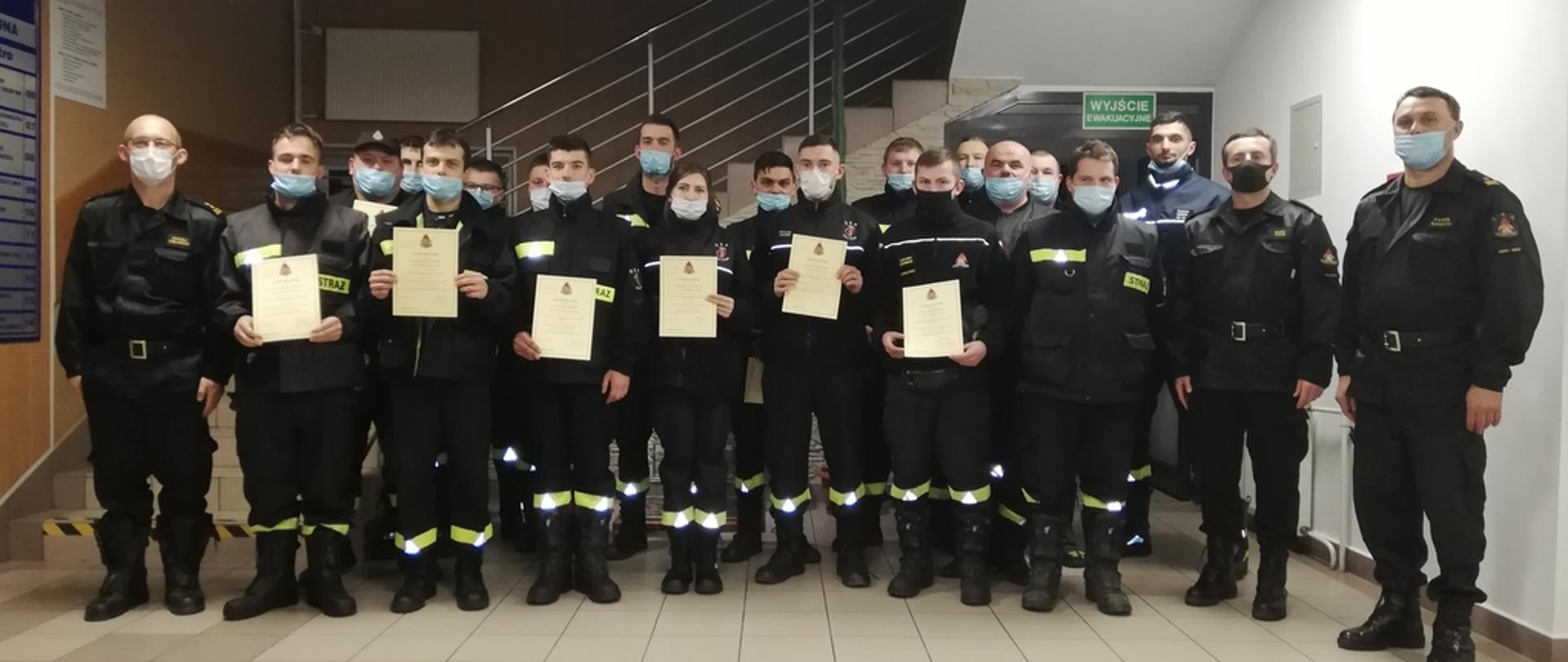 Na zdjęciu strażacy z Ochotniczych Straży Pożarnych po egzaminie kończącym szkolenie dowódców OSP. Strażacy ubrani w ubrania koszarowe koloru czarnego bez czapek trzymający w rękach zaświadczenia ukończenia szkolenie.
