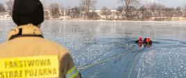 Strażacy samemu ewakuują się z przerębla na jednym z akwenów w Opolu. Strażacy w ubraniu z pianki wypornościowej oraz zabezpieczeni w kamizelki ratownicze, kaski i linę prowadzą ćwiczenia na akwenie z dziedziny ratownictwa wodnego. Na pierwszym planie strażak Państwowej Straży Pożarnej w umundurowaniu bojowym. W prawej części zdjęcia widoczne desko sanie ratownicze.