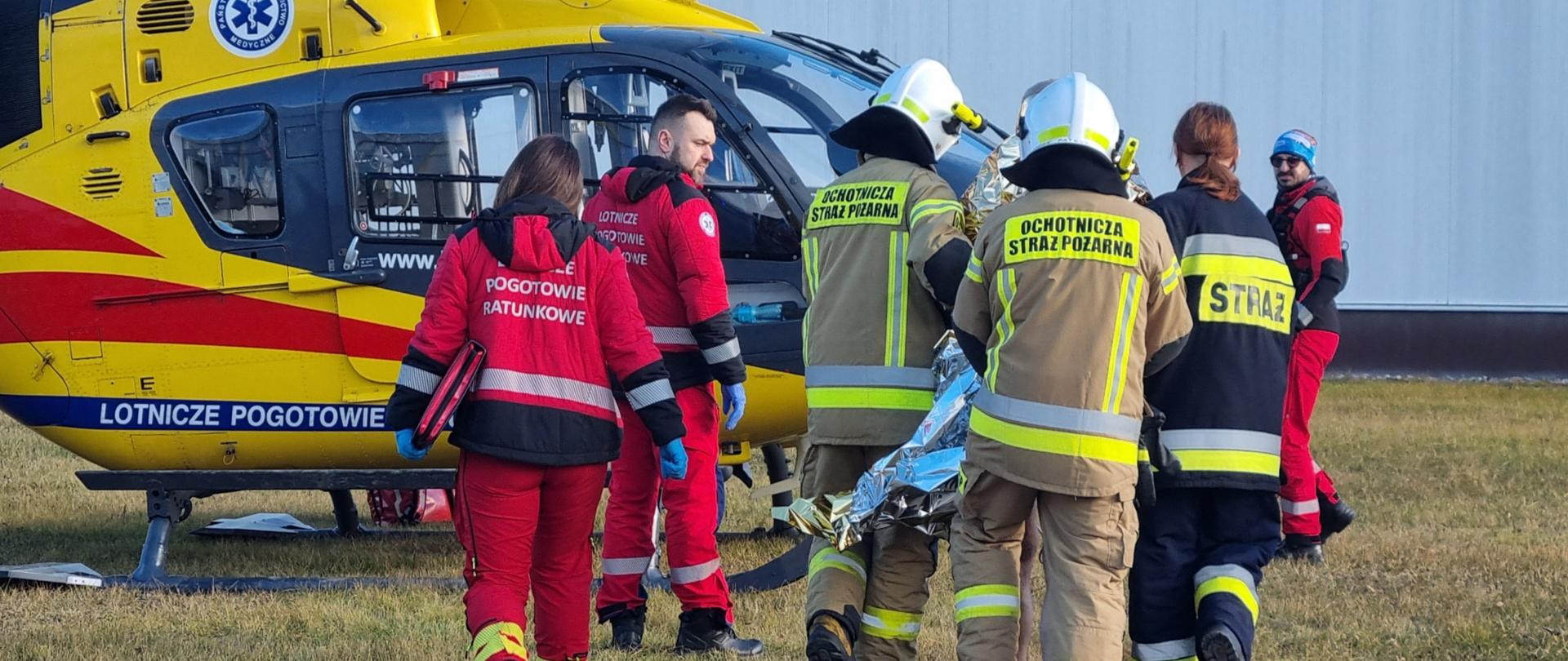 Strażacy wspólnie z ratownikami transportują osobę do śmigłowca Lotniczego Pogotowia Ratunkowego.