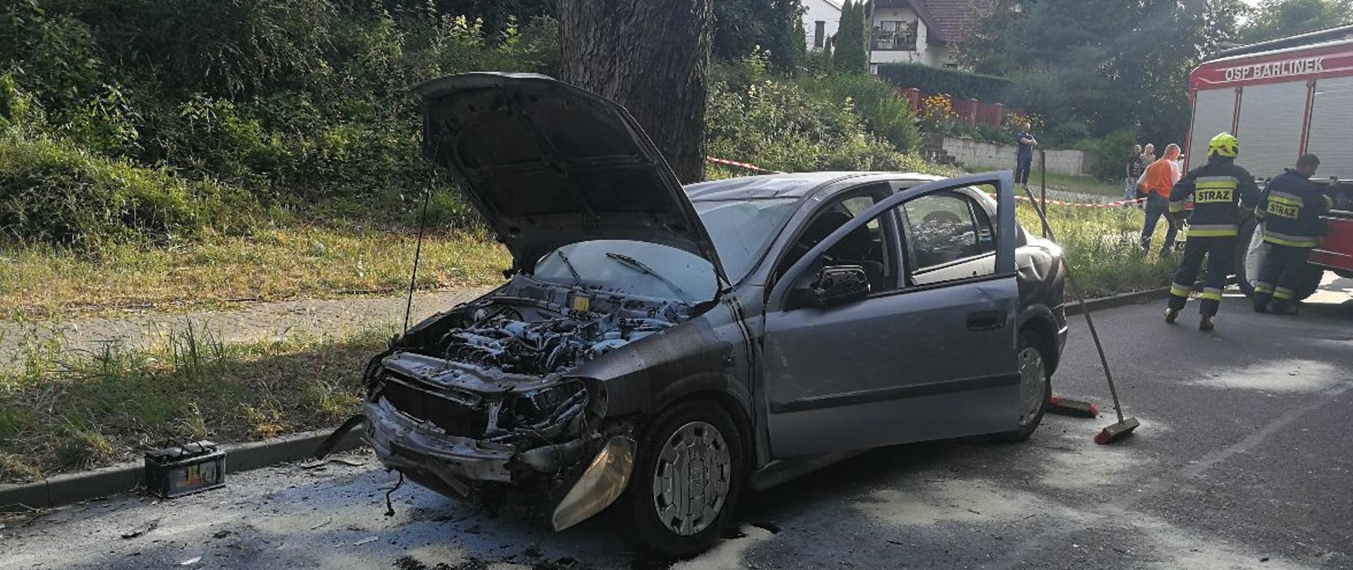 Na jezdni uszkodzony samochód osobowy. Wokół pracujący strażacy i pojazd pożarniczy.