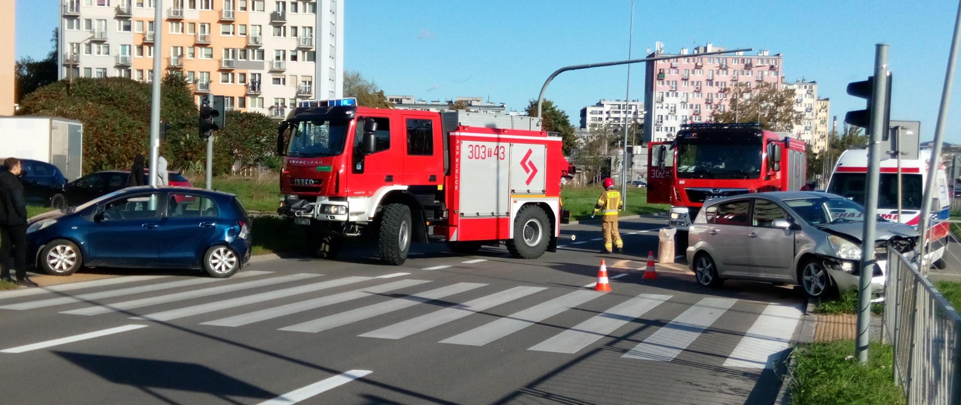 Zdjęcie przedstawia ulicę Grunwaldzką w Kielcach. Po dwóch stronach jezdni stoją auta biorące udział w zderzeniu. W głębi widać dwa samochody strażackie oraz ambulans. Między nimi przechodzi strażak, a na jezdni widać rozsypany sorbent zbierający wycieki. 