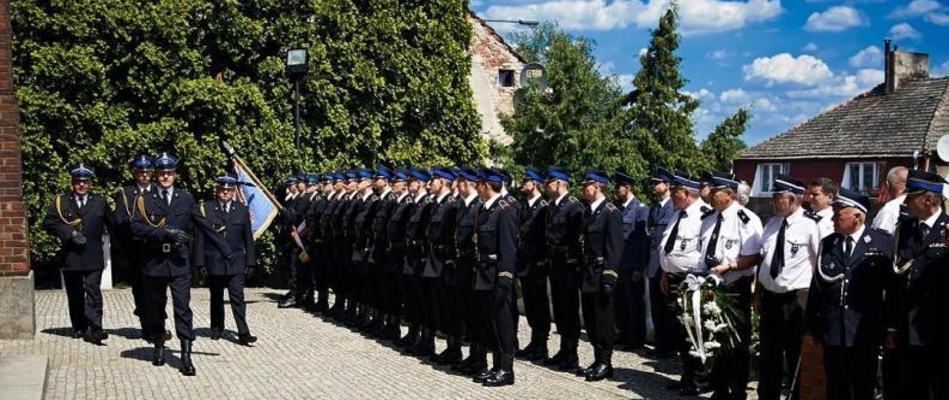 Na zdjęciu widzimy trzy szeregi mężczyzn strażaków ubranych w galowe mundury koloru czarnego, czapki na głowach, czarne buty. Przed szeregami strażaków maszeruje czterech strażaków, wchodzą do kościoła. W tle widzimy zielone drzewa, zabudowania mieszkalne, niebo koloru niebieskiego z białymi kłębami chmur. Strażacy stoją na kostce brukowej koloru szarego. 