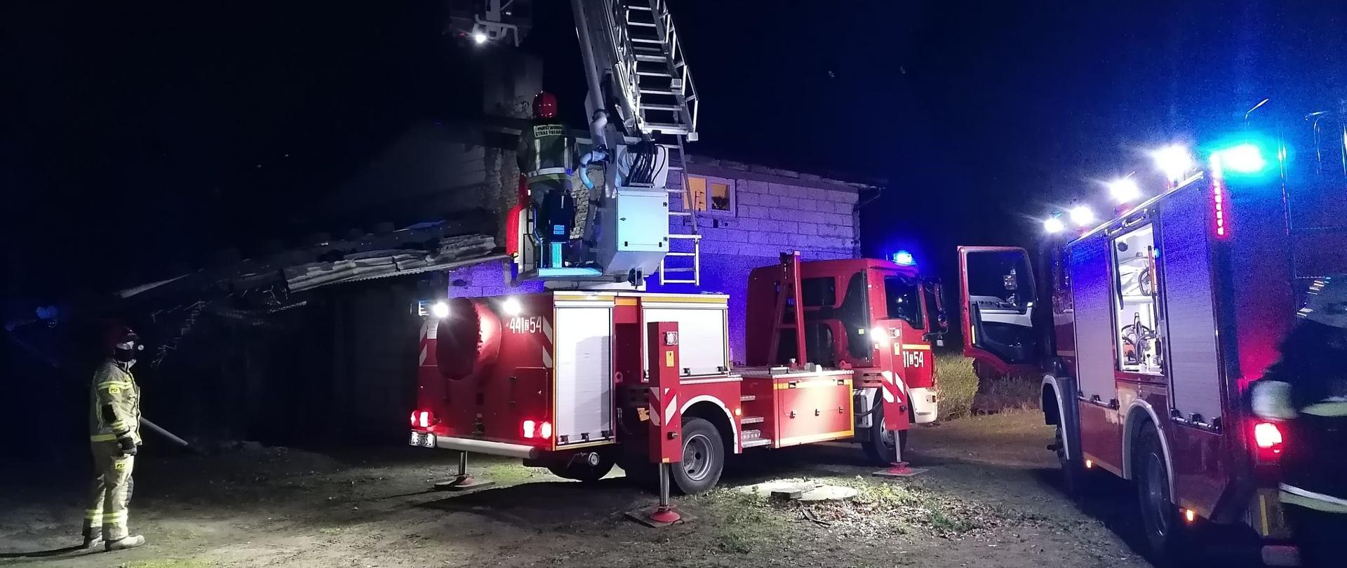 widoczny strażak oraz rozłożony podnośnik SHD-23 wraz z pojazdem gaśniczym na tle budynku mieszkalnego