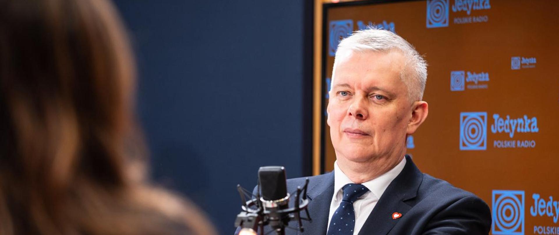 Zdjęcie przedstawia wnętrze studia radiowego. Po prawej stronie, siedzący za mikrofonem radiowym, minister Tomasz Siemoniak. Po lewej stronie widać sylwetkę siedzącej tyłem redaktor Agaty Kondzińskiej. Za ministrem pomarańczowa ścianka z białymi napisami i logiem "Jedynki". Tło lewej strony zdjęcia to granatowa ściana.