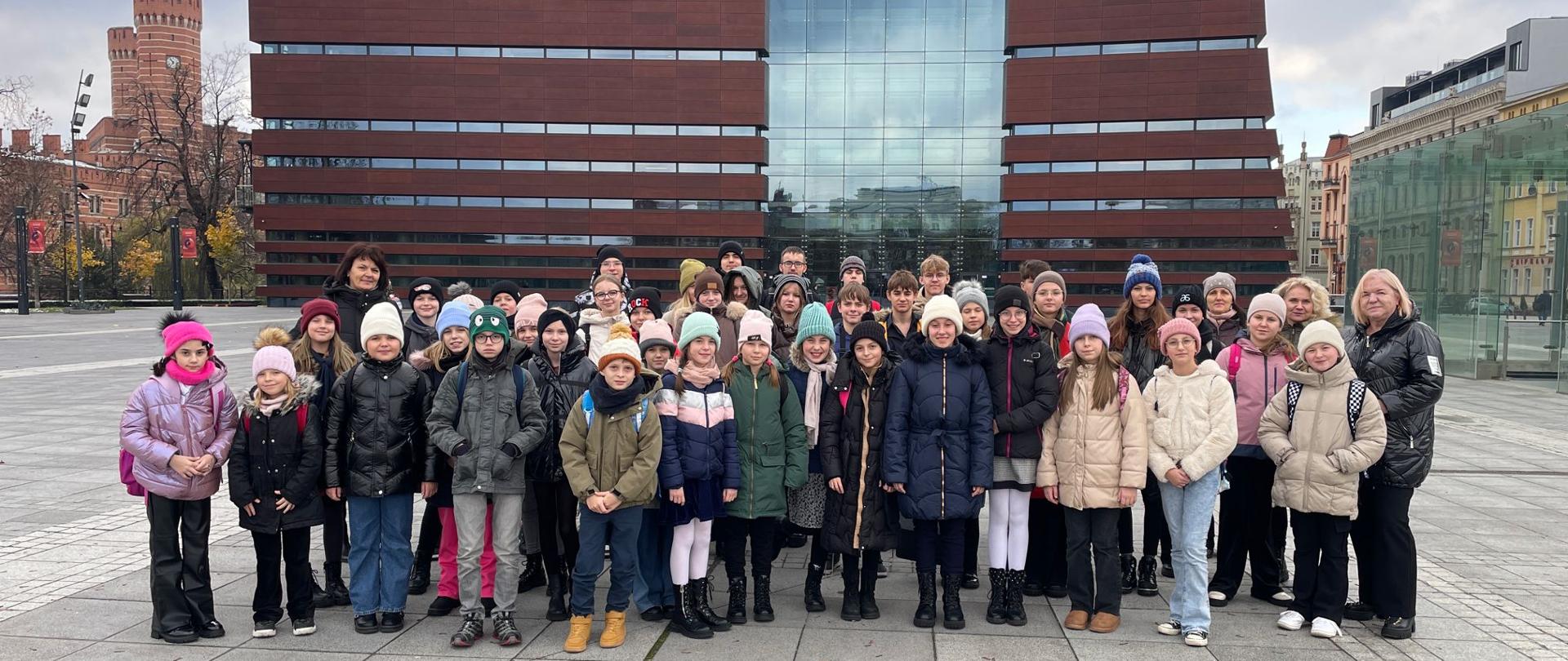 Zdjęcie grupowe uczniów z opiekunami na placu wyłożonym płytami przed gmachem Narodowego Forum Muzyki we Wrocławiu. Budynek w stylu nowoczesnym w bordowym kolorze z przeszkleniami.