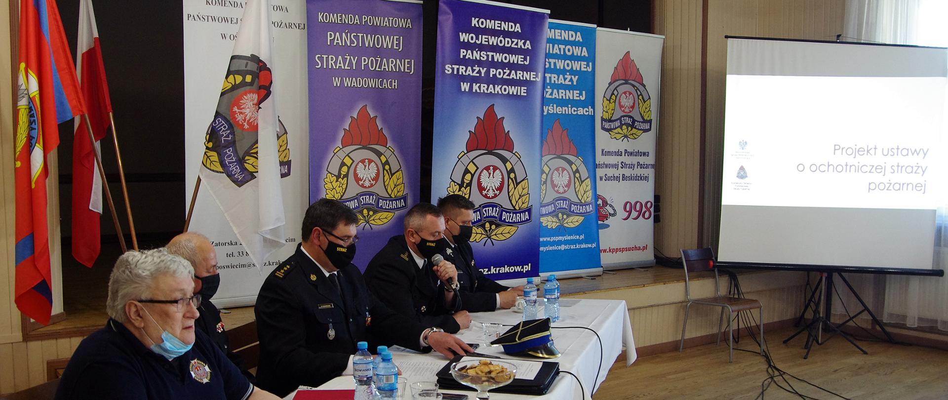 Widok z półprofilu. Na pierwszym widoczny stół prezydialny, za którym siedzą w mundurach strażackich i maseczkach ochronnych oficerowie PSP i członkowie OSP. W centralnej części siedzi Małopolski Komendant Wojewódzki PSP i omawia tekst projektu ustawy o OSP. W tle za stołem prezydialnym widoczne banery KW PSP Kraków i kilku KP PSP. Po prawej stronie ekran z tekstem projektu ustawy o PSP. 