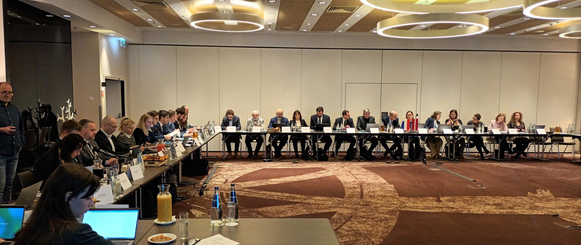 Sala konferencyjna. Uczestnicy konferencji (kobiety i mężczyźni) siedzą za stołem. Przed nimi laptopy i dokumentacja. 