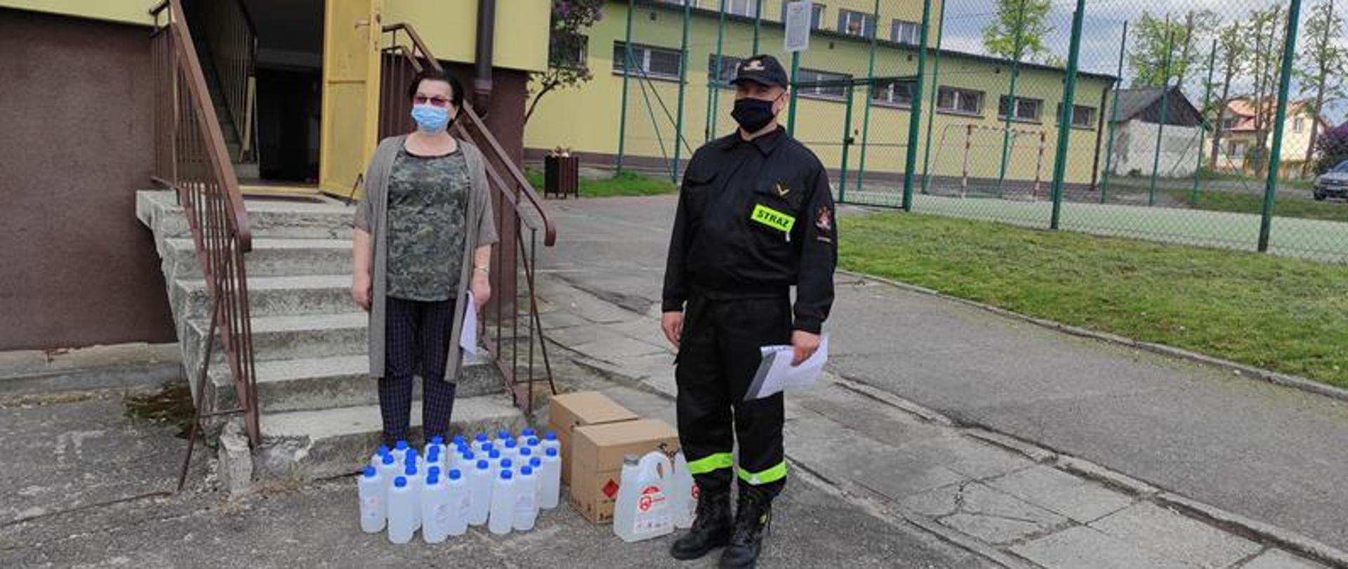 Strażak ubrany w maseczkę przekazuje przedstawicielowi szkoły podstawowej środki ochrony osobistej.