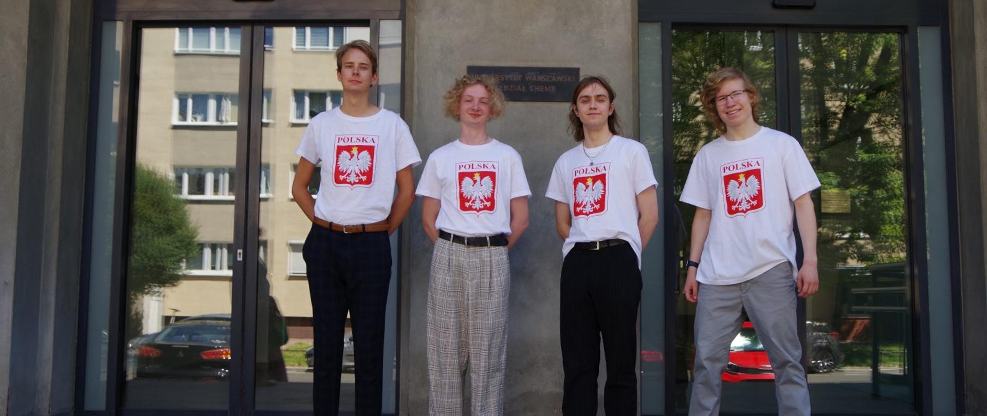 Czworo młodych ludzi w białych koszulkach z godłem Polski stoi przed ponuro wyglądającym, brudnoszarym budynkiem.