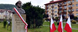 Na zdjęciu widać pomnik Generała Stanisława Maczka obok którego stoją 3 flagi Polski. W tle widać blok oraz dworzec. 