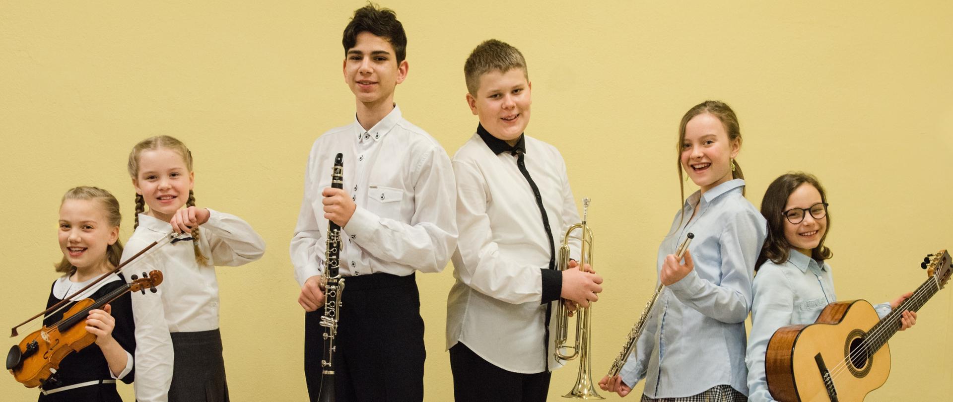 Zdjęcie przedstawia sześcioro uczniów na tle żółtej ściany (4 dziewczynki, 2 chłopców), trzymających instrumenty muzyczne (skrzypce, klarnet, trąbka, flet, gitara)