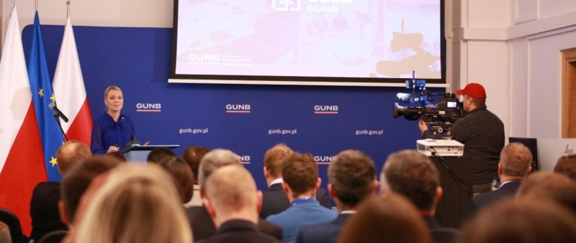 Prelegent przemawia na tle ścianki GUNB, obok flag Polski i UE, podczas konferencji z udziałem publiczności.