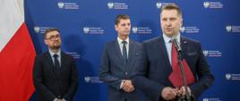 Minister Czarnek stoi i mówi do mikrofonu na stojaku, za nim wiceminister Piontkowski i mężczyzna w okularach, za nimi niebieska ściana i polska flaga.