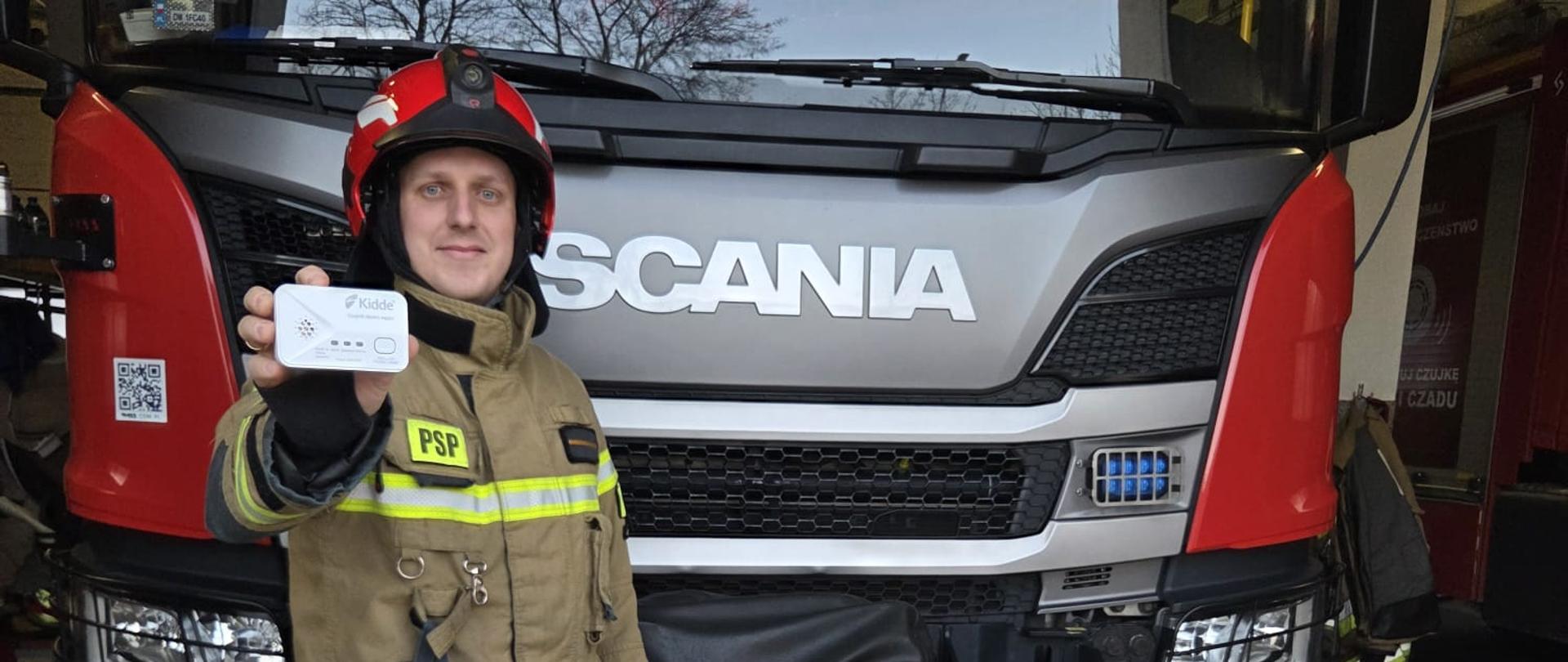 Strażak w pełnym umundurowaniu bojowym stoi przed frontem wozu strażackiego marki Scania i trzyma w wyciągniętej dłoni czujkę tlenku węgla marki Kidde. Strażak ma na sobie piaskowy kombinezon z napisem „PSP”, czerwony hełm oraz rękawice przypięte do pasa. Zdjęcie zostało wykonane w garażu remizy, a jego celem jest promocja bezpieczeństwa i stosowania czujek czadu.