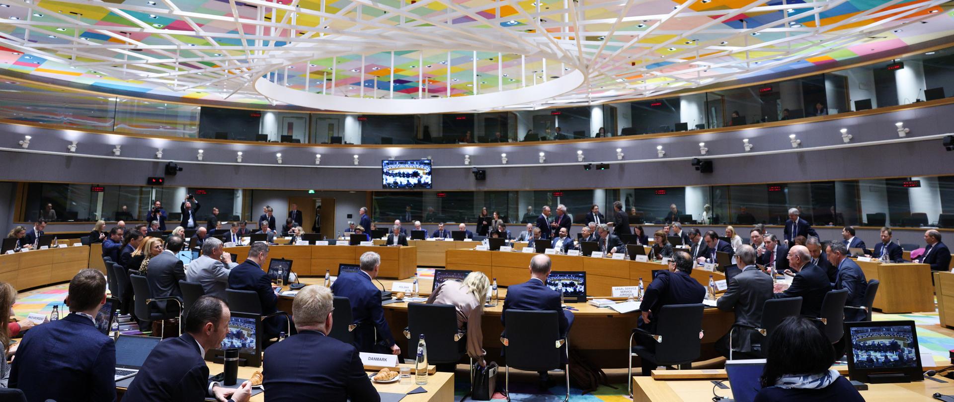 Sala obrad z uczestnikami posiedzenia ECOFIN przy okrągłym stole, pod kolorowym, geometrycznym sufitem z oświetleniem