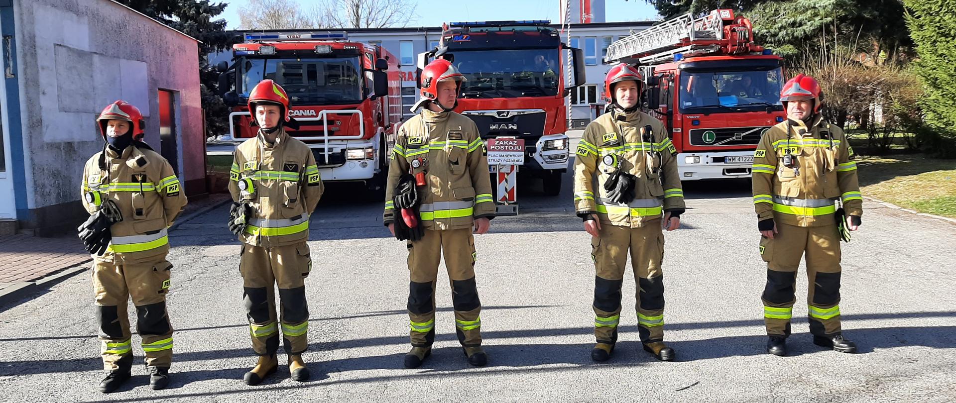 Strażacy stoją na placu przez Komendą, w tle samochody pożarnicze z włączonymi sygnałami świetlnymi i dźwiękowymi
