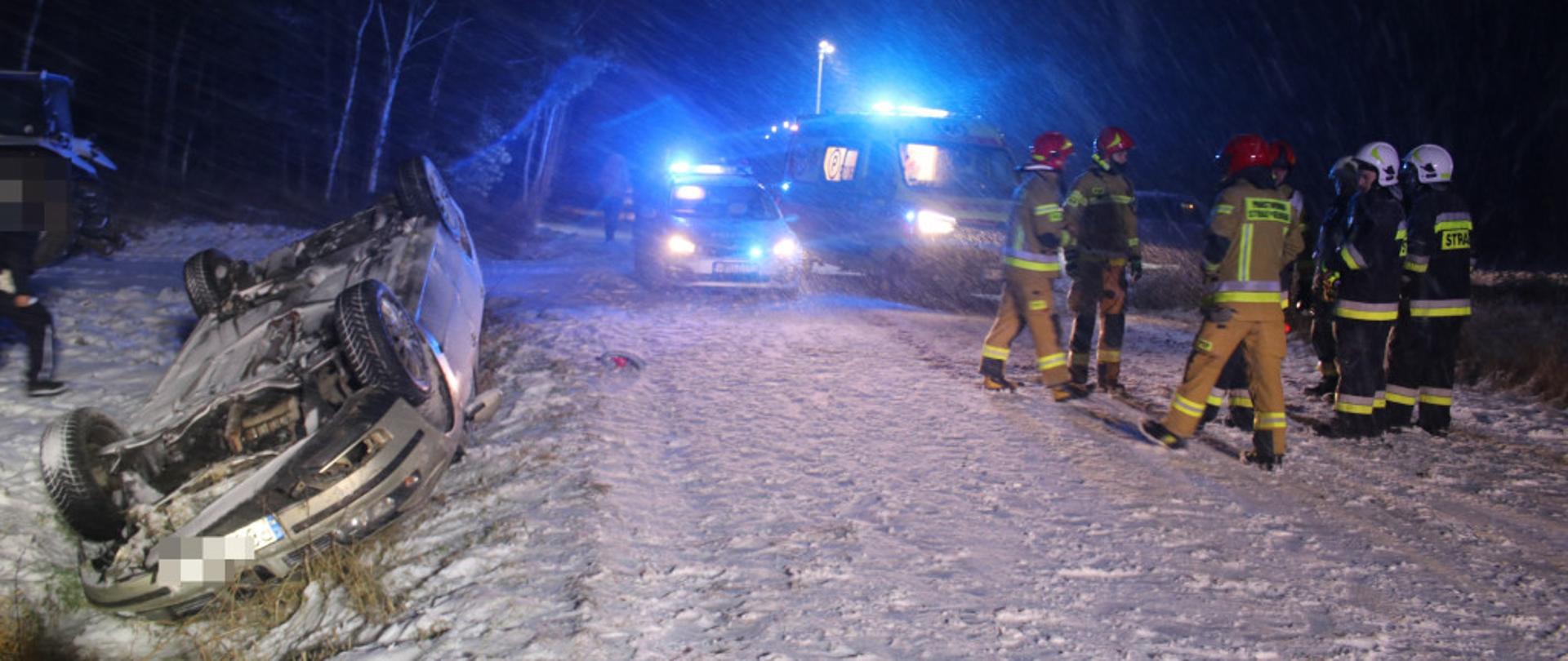 Zdjęcie przedstawia samochód osobowy leżący na dachu. W tle stoją strażacy.