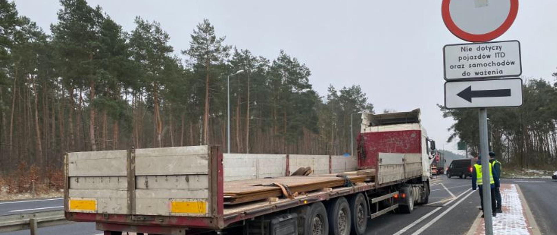 Ciężarówka z naczepą. Burta naczepy jest otwarta. Obok stoją inspektorzy. Na naczepie metalowe płyty.