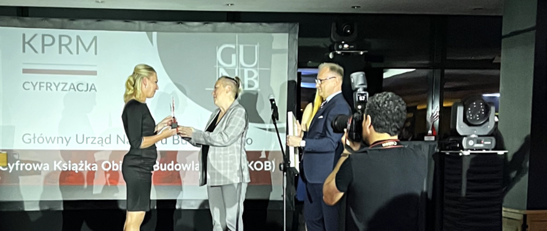 Trzy osoby na scenie podczas ceremonii wręczenia nagrody; w tle ekran z napisem „Cyfrowa Książka Obiektu Budowlanego (c-KOB)”.
