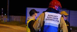Na przejeżdzie kolejowym stoi 2 strażaków i policjant. Strażak odwrócony tyłem ma na sobie biało-niebieską kamizelkę z napisem KIERUJĄCY DZIAŁANIEM RATOWNICZYM. Pora nocna.