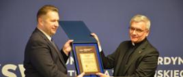 Minister Czarnek i mężczyzna w czarnym garniturze z koloratką trzymają razem otwarte płaskie pudełko, pokazują jego zawartość - wygląda jak ozdobny dyplom leżący na niebieskim pluszu.