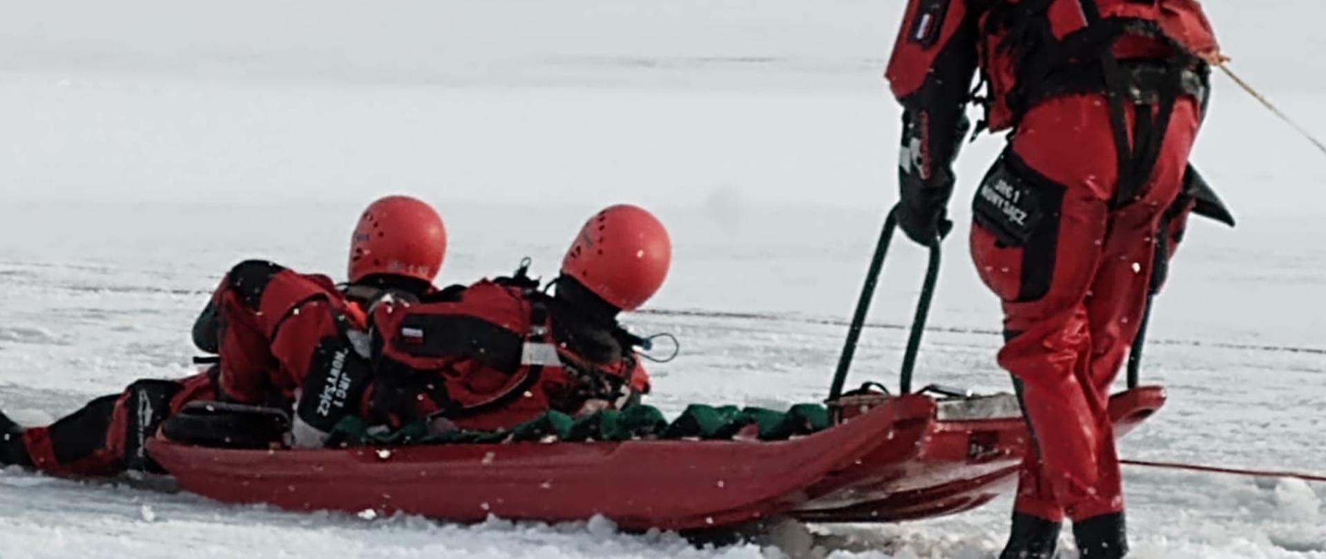 Dwóch strażaków na saniach lodowych Trzeci je ciągnie za uchwyty.