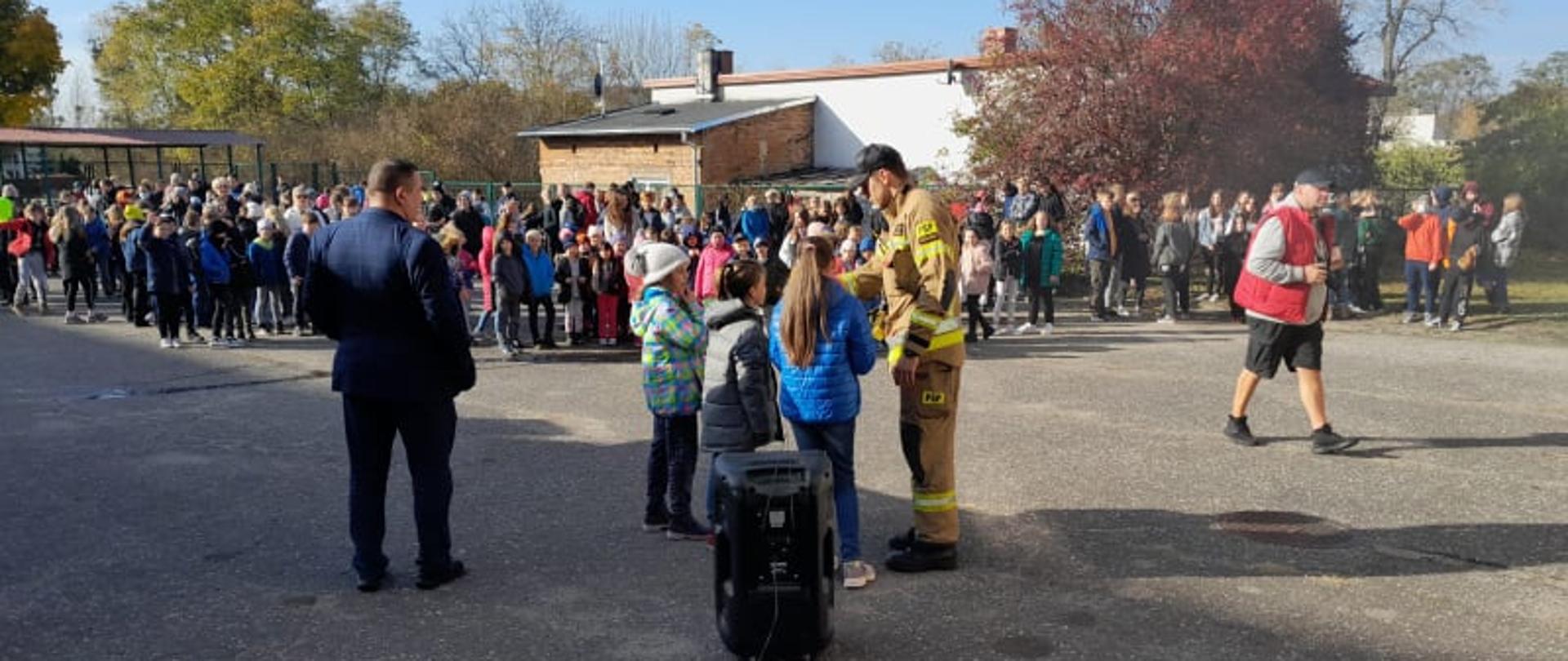 Na zdjęciu widać trójkę dzieci oraz strażaka podczas udzielania odpowiedzi na pytania. Obok stoi dyrektor szkoły. Wszyscy zwróceni są w kierunku dzieci ustawionych po ewakuacji. Całość ma miejsce na placu szkolnym w tle widać zabudowania jednorodzinne oraz drzewa w jesiennych barwach.
