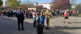 Na zdjęciu widać trójkę dzieci oraz strażaka podczas udzielania odpowiedzi na pytania. Obok stoi dyrektor szkoły. Wszyscy zwróceni są w kierunku dzieci ustawionych po ewakuacji. Całość ma miejsce na placu szkolnym w tle widać zabudowania jednorodzinne oraz drzewa w jesiennych barwach.
