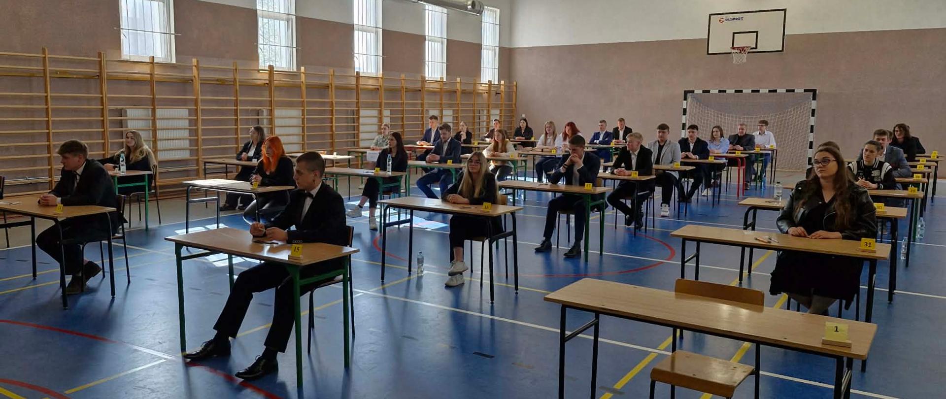 Uczniowie siedzący przy numerowanych stolikach w sali gimnastycznej.