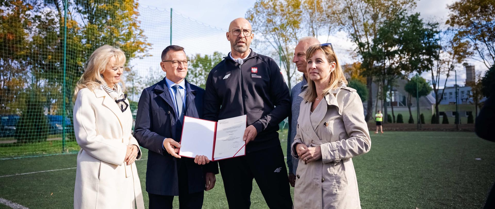 Zdjęcie przedstawiające uczestników konferencji w Żninie stojących na Orliku. Na środku stoją minister sportu i turystyki Jakub Rutnicki i burmistrz Żnina Łukasz Kwiatkowski trzymając decyzję o przyznaniu dofinansowania na remont Orlika.