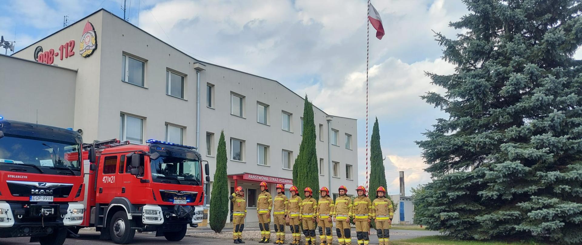 Zdjęcie przed Komendą Powiatową PSP w Opocznie przedstawia dwa wozy bojowe z włączonymi sygnałami świetlnymi oraz strażaków w umundurowaniu specjalnym oddających honory.