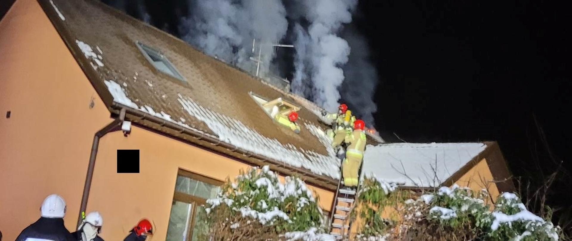 Strażacy pracujący na dachu budynku mieszkalnego, gdzie powstał pożar. Są zabezpieczeni linkami ratowniczymi przy pracach po pożarze. 