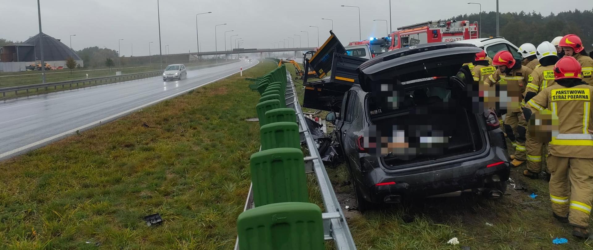 Na zdjęciu z prawej strony widać rozbity samochód osobowy stojący na pasie zieleni przy barierkach na środku autostrady. Pojazd ma otwarta klapę bagażnika. Obok stoją strażacy w żółtych strojach oraz białych i czerwonych hełmach. W tle widać rozbity samochód dostawczy oraz czerwone samochody strażackie. Po lewej stronie zdjęcia widać jezdnie na drugim pasie autostrady, po której jedzie srebrny samochód. W tle widać budynki oraz wiadukt. Jest widno, ale pochmurnie. 