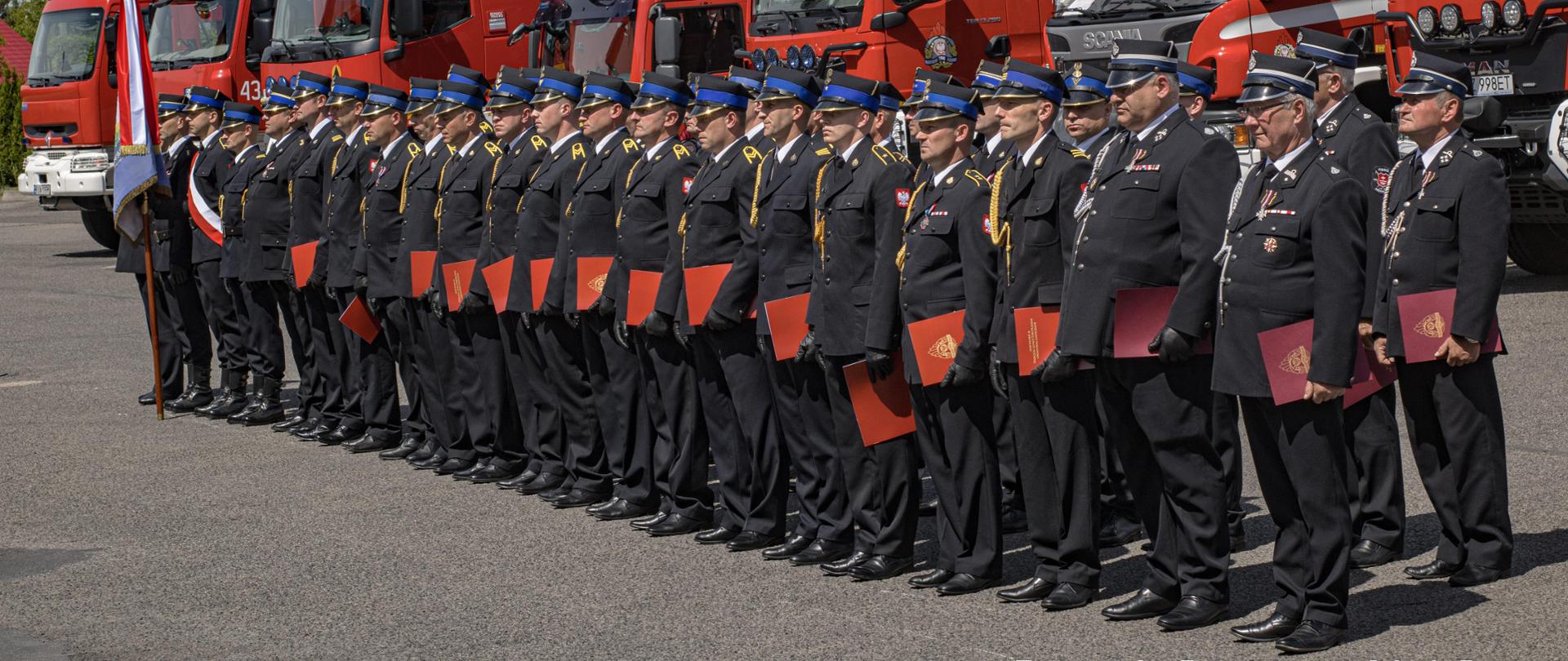 Zdjęcie przedstawia pododdział strażaków na czele z pocztem sztandarowym, ustawiony w dwuszeregu na placu wewnętrznym komendy. Strażacy ubrani w umundurowanie wyjściowe, niektórzy trzymają w lewej długi akty mianowania na wyższy stopień służbowy. W tle widoczne są ustawione w szeregu pojazdy pożarnicze.